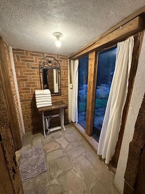 A bathroom with a stone floor, a bench with a towel, a mirror, and a window with curtains. - Cabañas del Valle  (San Pedro Atmatla)