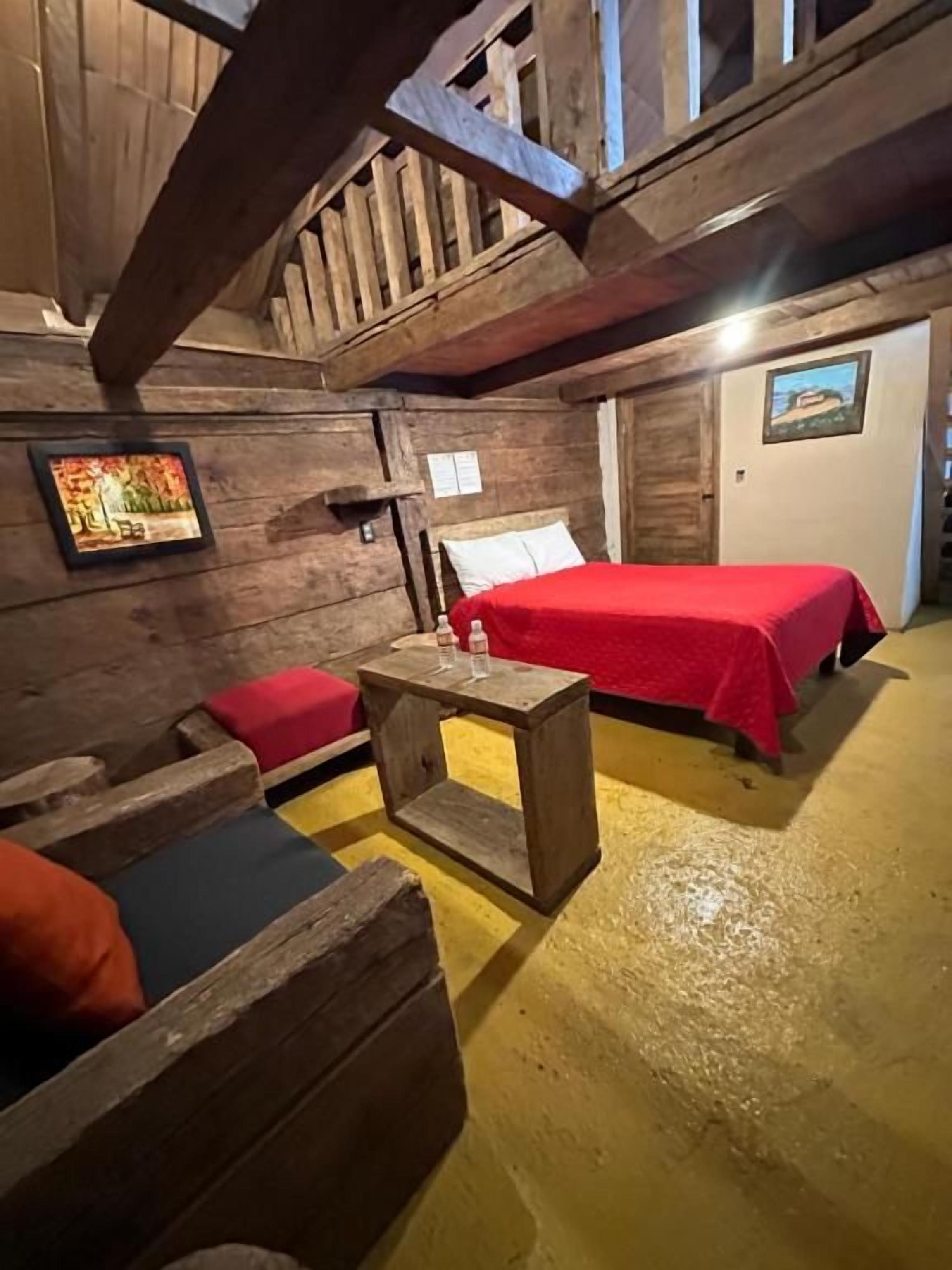 Una habitación con paredes de madera y techo inclinado, con dos camas con ropa roja, una mesita de madera y un banco con cojines.