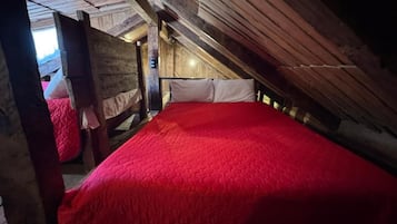 A room with a sloped ceiling, wooden beams, a red bedspread, and a window allowing natural light.