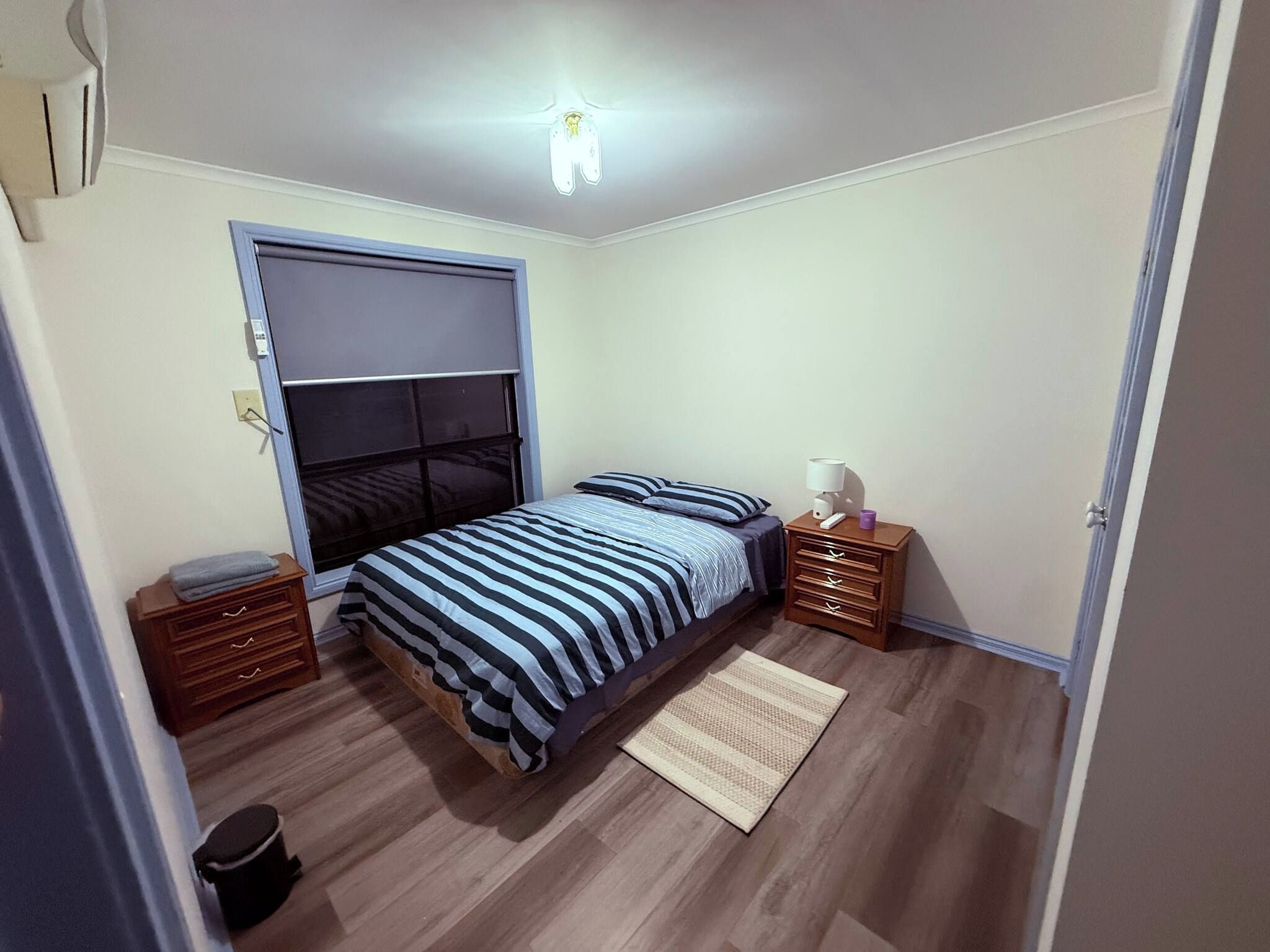 Et soveværelse med seng, natbord, kommode, et vindue med persienner og en loftlampe.