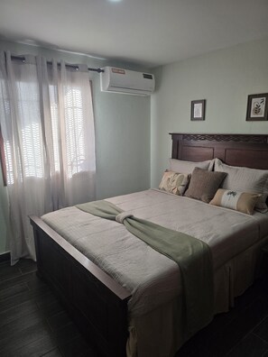 A bedroom with a bed, a headboard, a window with curtains, and an air conditioning unit. - HISTORIAL COLONIAL ZONE UNIT, LARGE TERRACE, VIEW OF THE CARIBBEAN SEA (Santo Domingo)