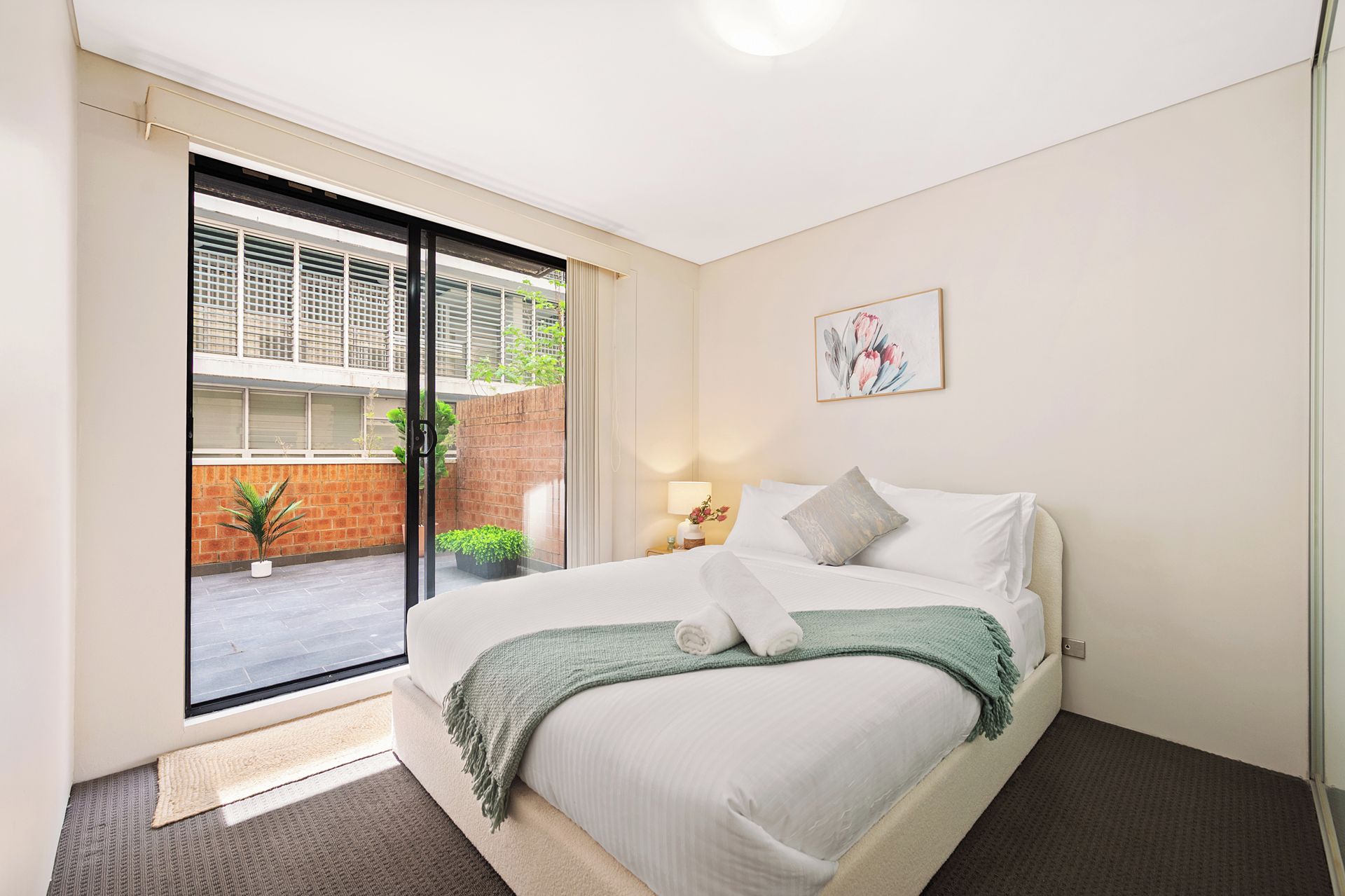 A hotel room with a bed, a window, a door, and a small table with a lamp.