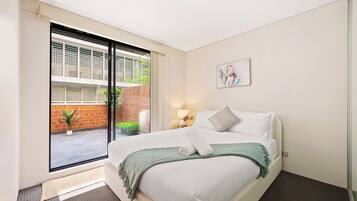 A hotel room with a bed, a window, a door, and a small table with a lamp.