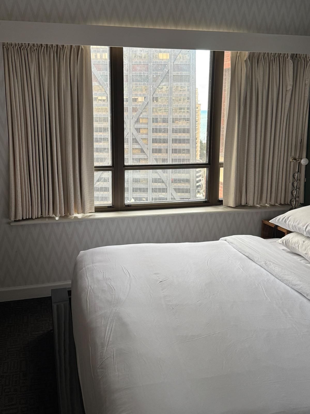 Habitación de hotel con una cama grande, una ventana con cortinas y vistas a un edificio.