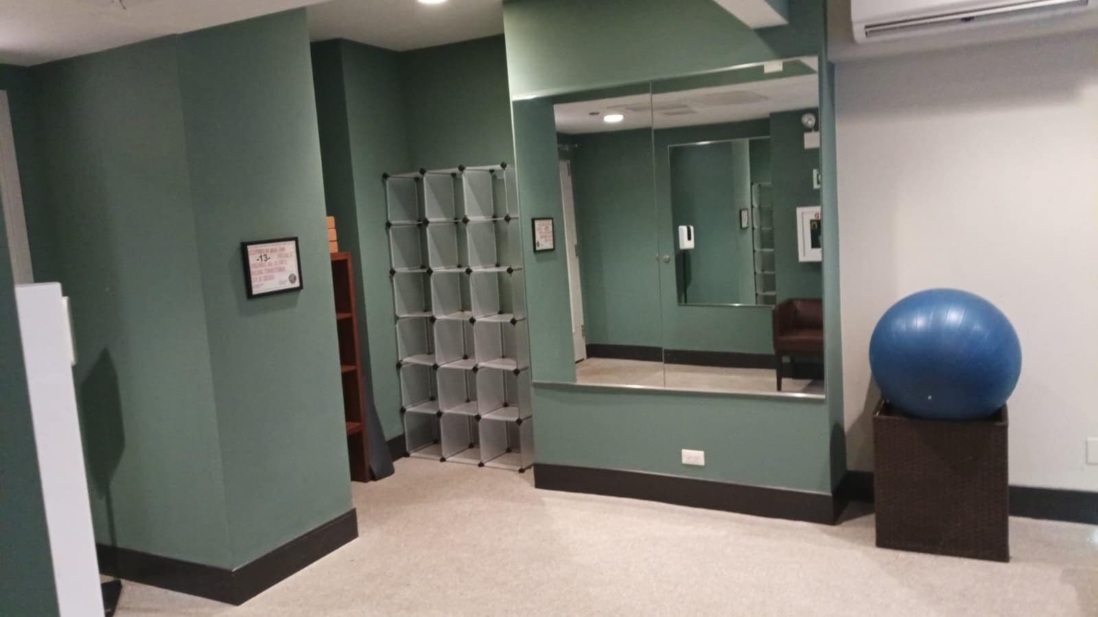 Una habitación con paredes verde azulado, un armario con espejo, una pelota de ejercicios azul sobre un soporte y un certificado enmarcado en la pared.