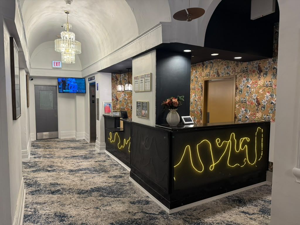 Zona de recepción de un hotel con un mostrador negro, una araña de cristal, un televisor y un papel tapiz con motivos florales.