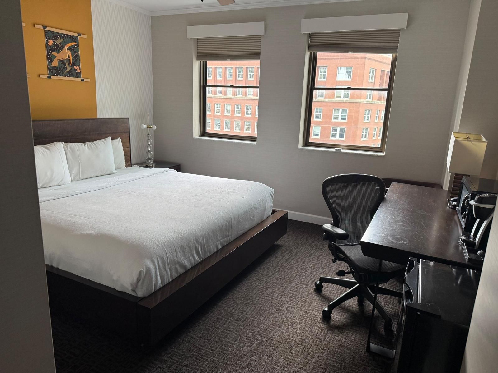 Een hotelkamer met een bed, een bureau, een stoel en twee ramen met uitzicht op gebouwen.
