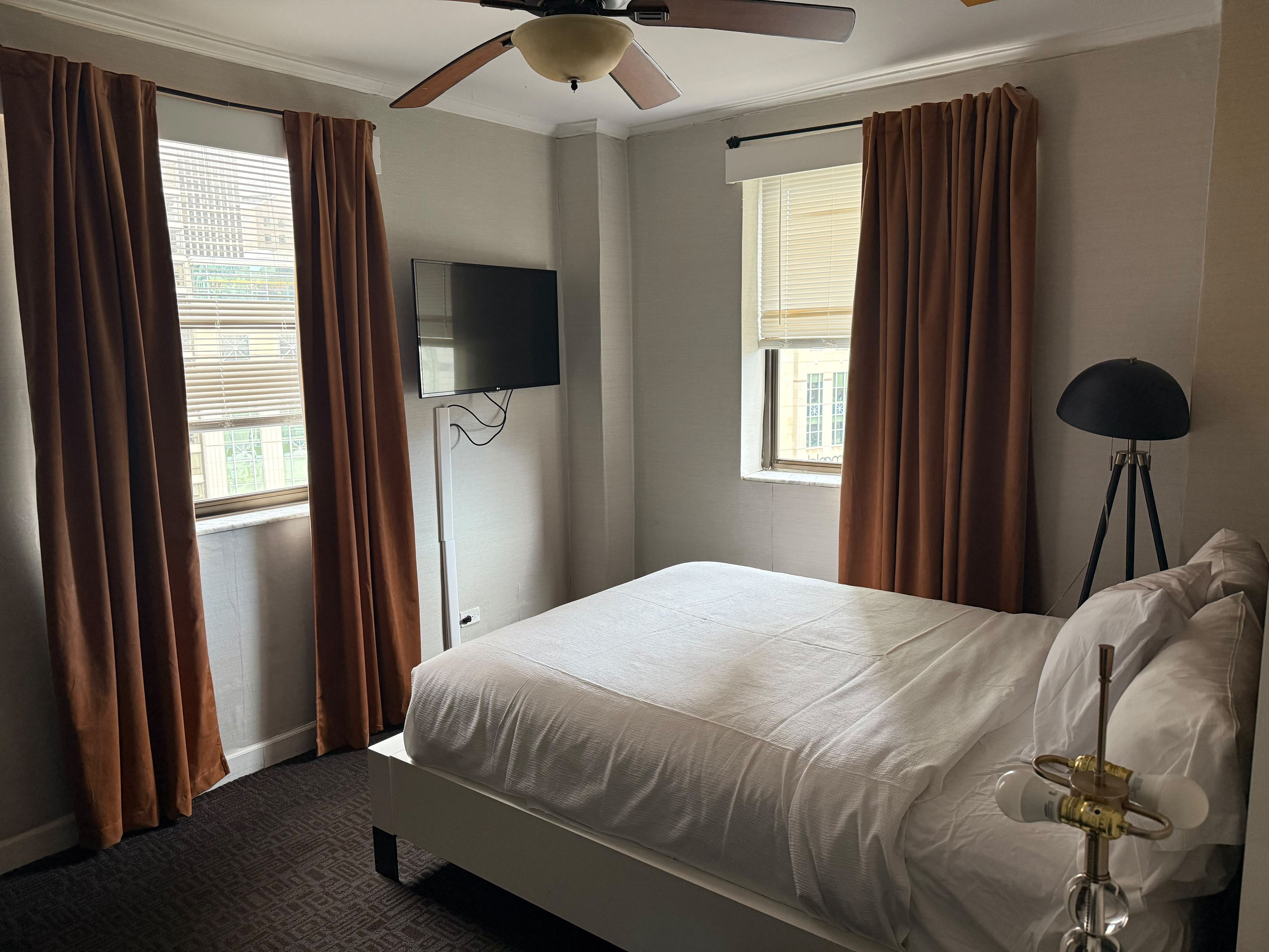 Een hotelkamer met een bed, een plafondventilator, een aan de muur bevestigde televisie en een vloerlamp.