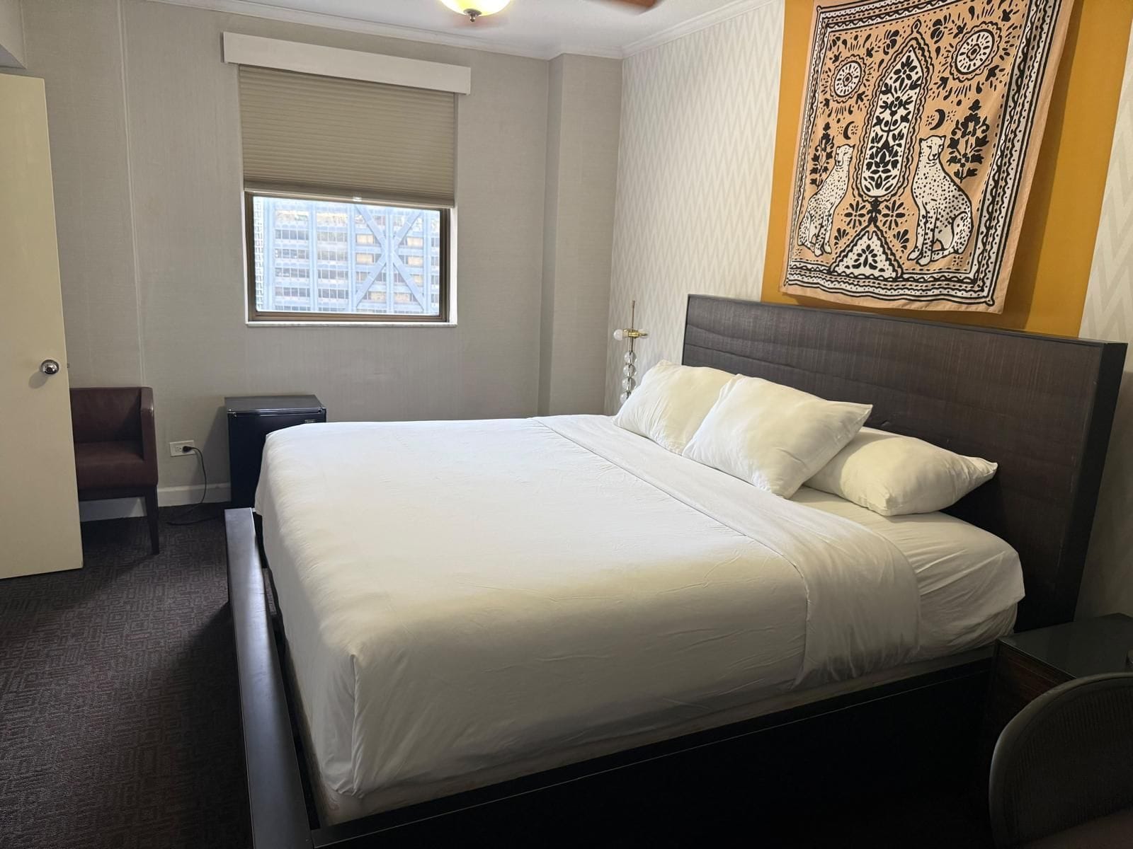 Ein Hotelzimmer mit einem großen Bett, einem Fenster mit Jalousien, einem Sessel, einem Schreibtisch und einem Wandteppich.