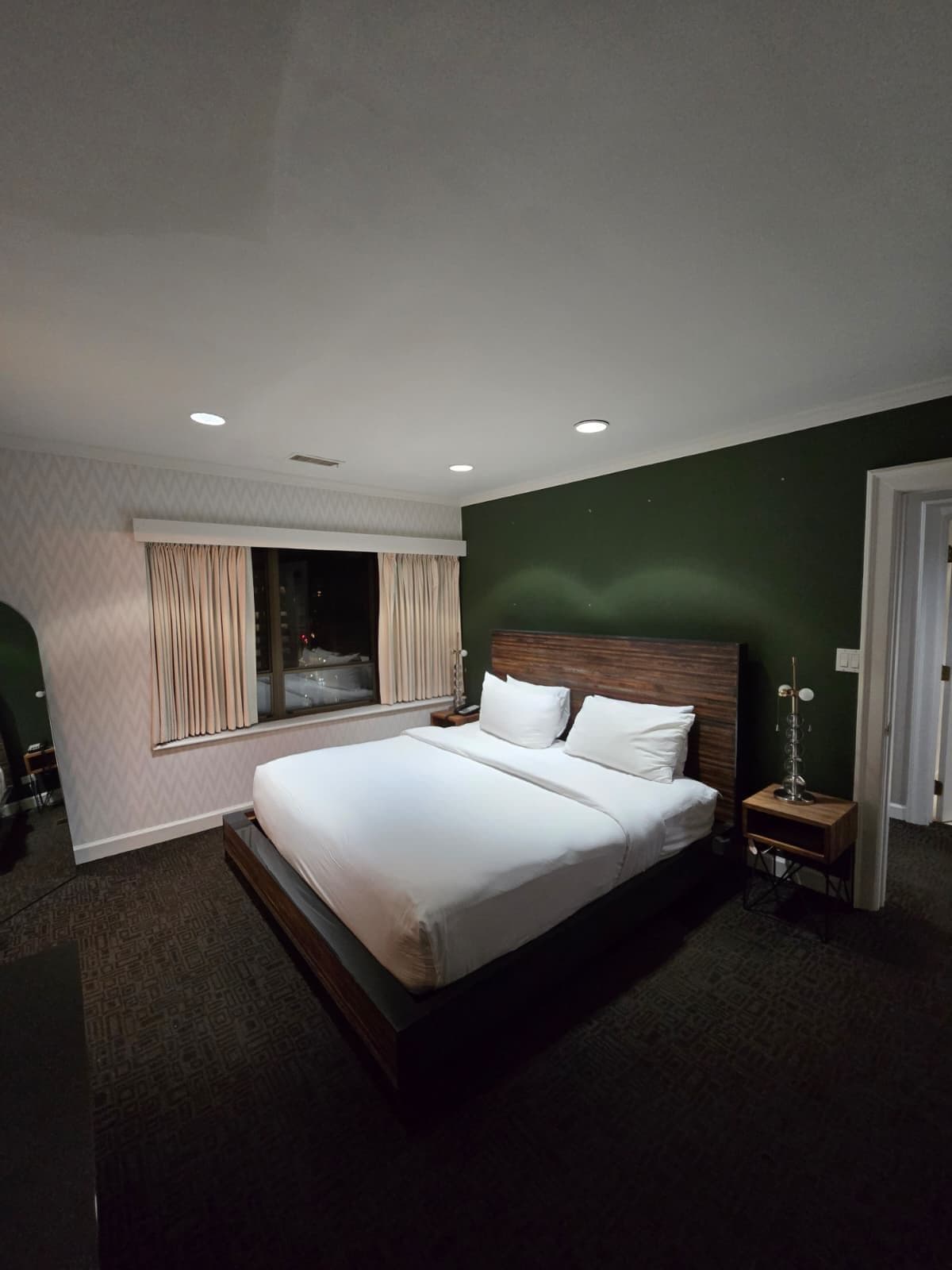 A modern hotel room with a large bed, a bedside table, a nightstand, and a window with curtains.