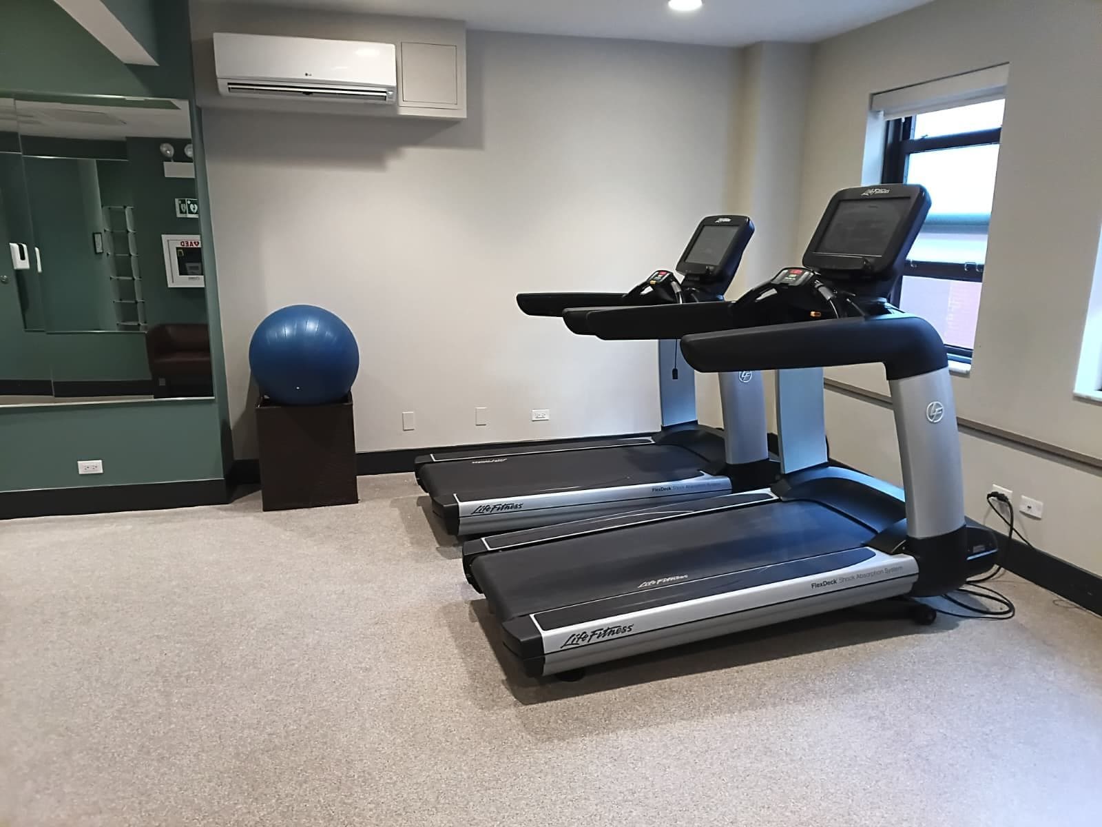 Un gimnasio con cintas de correr, una pelota de ejercicios azul y un equipo de aire acondicionado instalado en la pared.