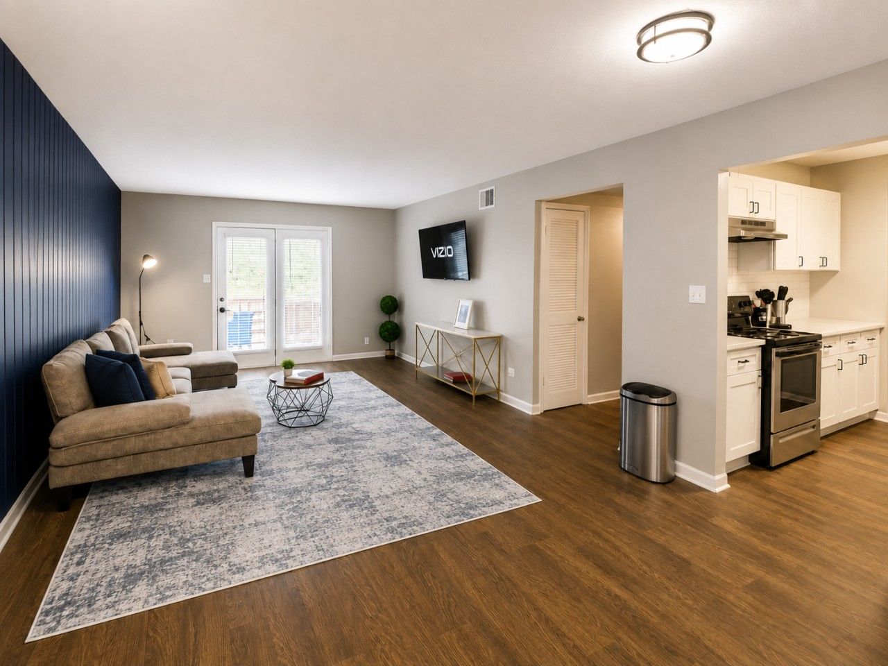 A modern living room with a sofa, a coffee table, and a rug. There is a kitchen area with cabinets and a stove.