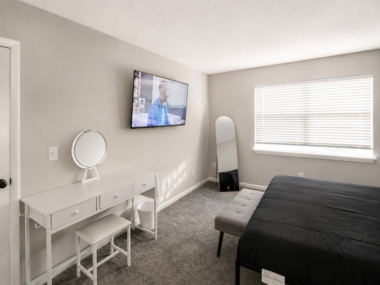 A bedroom with a bed, a desk with a mirror, a television, and a window with blinds.