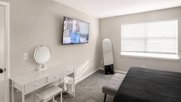 A bedroom with a bed, a desk with a mirror, a television, and a window with blinds.