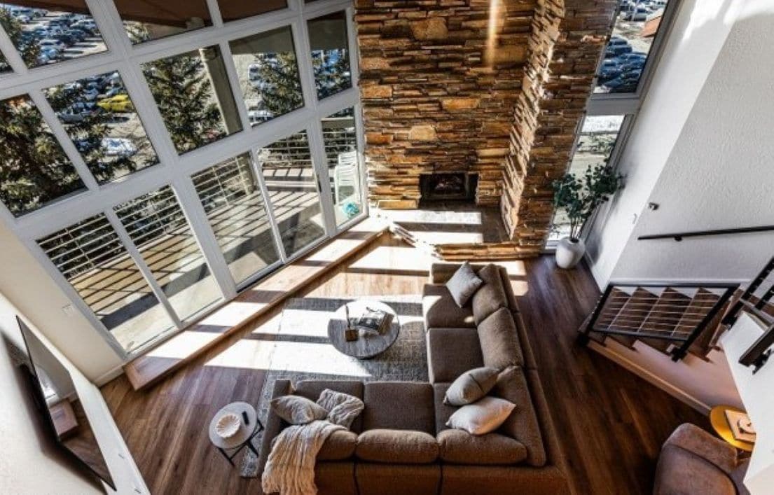 A modern living room with a stone fireplace, large windows, and a wooden floor.