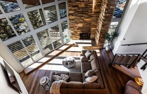 A modern living room with a stone fireplace, large windows, and a wooden floor. - IL CERVO - The Stag (Park City)