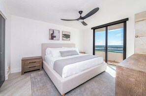 Une chambre à coucher avec un lit, des tables de chevet, un ventilateur de plafond, des œuvres d’art accrochées au mur et une vue dégagée sur l’extérieur grâce à une grande fenêtre.