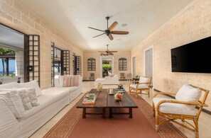 A spacious living area with a sofa, coffee table, and wicker chairs. A ceiling fan and a flat-screen TV are mounted on the wall. - L'Horizon Luxury Sea View Villa (Bridgetown)