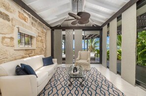 A modern living room with a stone wall, a ceiling fan, a white sofa, a patterned rug, and a view of the outdoors. - L'Horizon Luxury Sea View Villa (Bridgetown)