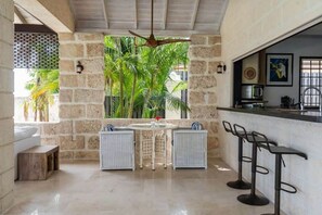 A kitchen with a stone wall, a ceiling fan, a window with a view of palm trees, two bar stools, and a countertop with kitchen appliances. - L'Horizon Luxury Sea View Villa (Bridgetown)