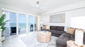 A spacious living room with a large sofa, a round coffee table, and a view of the ocean through the sliding glass doors. - #704 Sanibel Oceanfront 4/3 Condo. (Daytona Beach Shores)