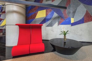 Un hall d’entrée moderne avec un espace détente aux fauteuils rouges, une table noire et un mur décoré de motifs géométriques colorés.
