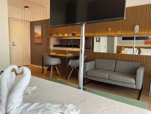 Habitació d'hotel amb un llit, un sofà, una taula de treball amb cadires, una televisió i una zona de cuina.