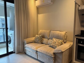 Ein Sofa mit gemusterten Kissen in einem Zimmer mit einem großen Fenster, einer Klimaanlage und einem Küchenbereich.