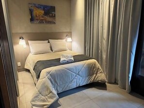 Ein Hotelzimmer mit Bett, zwei Nachttischlampen, einem Bild an der Wand und einem Fenster mit Vorhängen.
