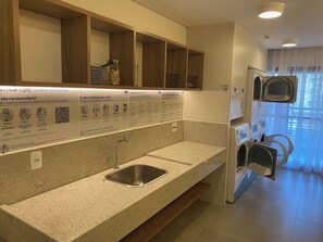 A hotel bathroom with a granite countertop, a sink, and a mirror. There are multiple towel dispensers and a wall-mounted washing machine. - Brand-New Studio near Brooklin Metro HMB616 (São Paulo)