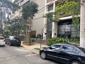 A street view with a black car parked on the side, a white car in the background, and a building with a modern design and a staircase leading to the entrance. - Brand-New Studio near Brooklin Metro HMB616 (São Paulo)