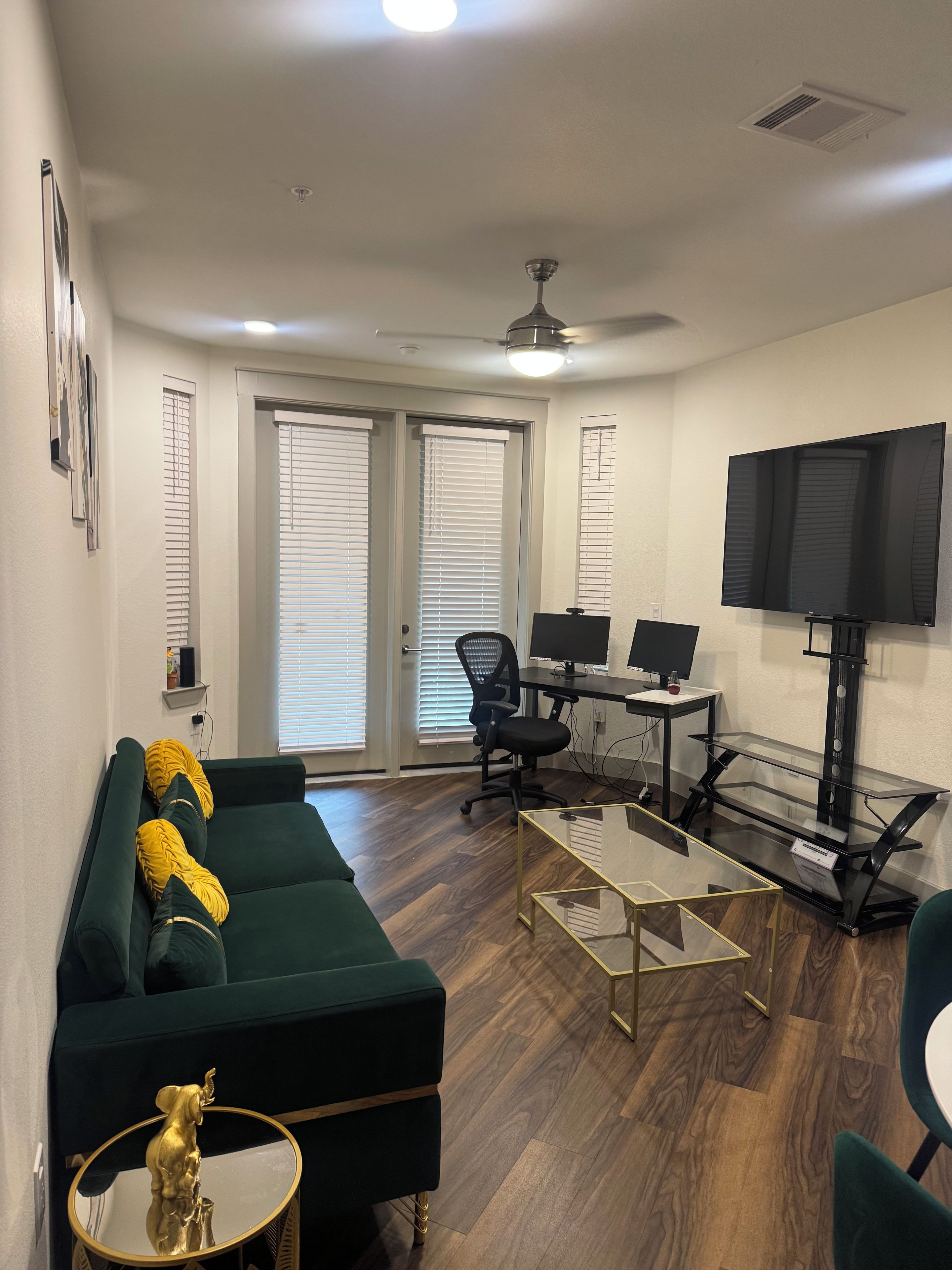 A living room with a green sofa, a glass coffee table, a black office chair, a TV on a stand, and a ceiling fan.