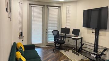 A living room with a green sofa, a glass coffee table, a black office chair, a TV on a stand, and a ceiling fan.