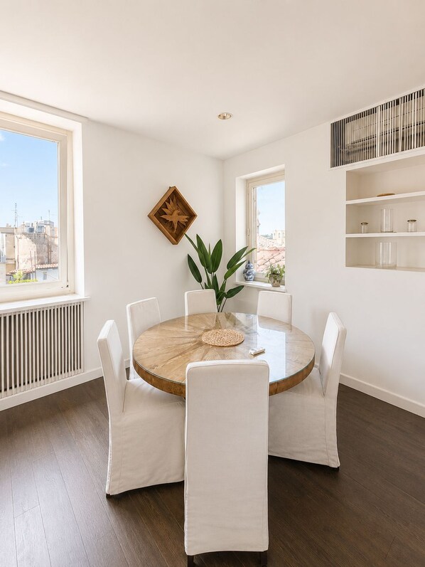 Espaço de refeições com mesa redonda, cadeiras brancas, planta em vaso e vista para a cidade através de amplas janelas.