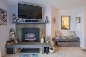 A living room with a fireplace, a flat-screen TV, a sofa, and a bench. - Stylish Top-Floor Ski-In/Out | Okemo Base Lodge (Ludlow)