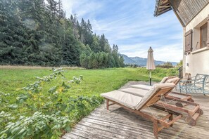 Une terrasse dotée de chaises longues et d’une table, offrant une vue sur une zone verdoyante avec des arbres et une montagne en arrière-plan.