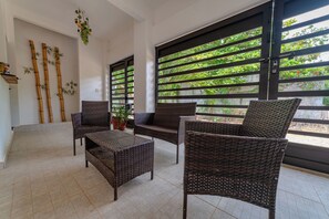 Una habitación con mobiliario de mimbre, una planta en maceta y vistas a través de una ventana con celosías de bambú.