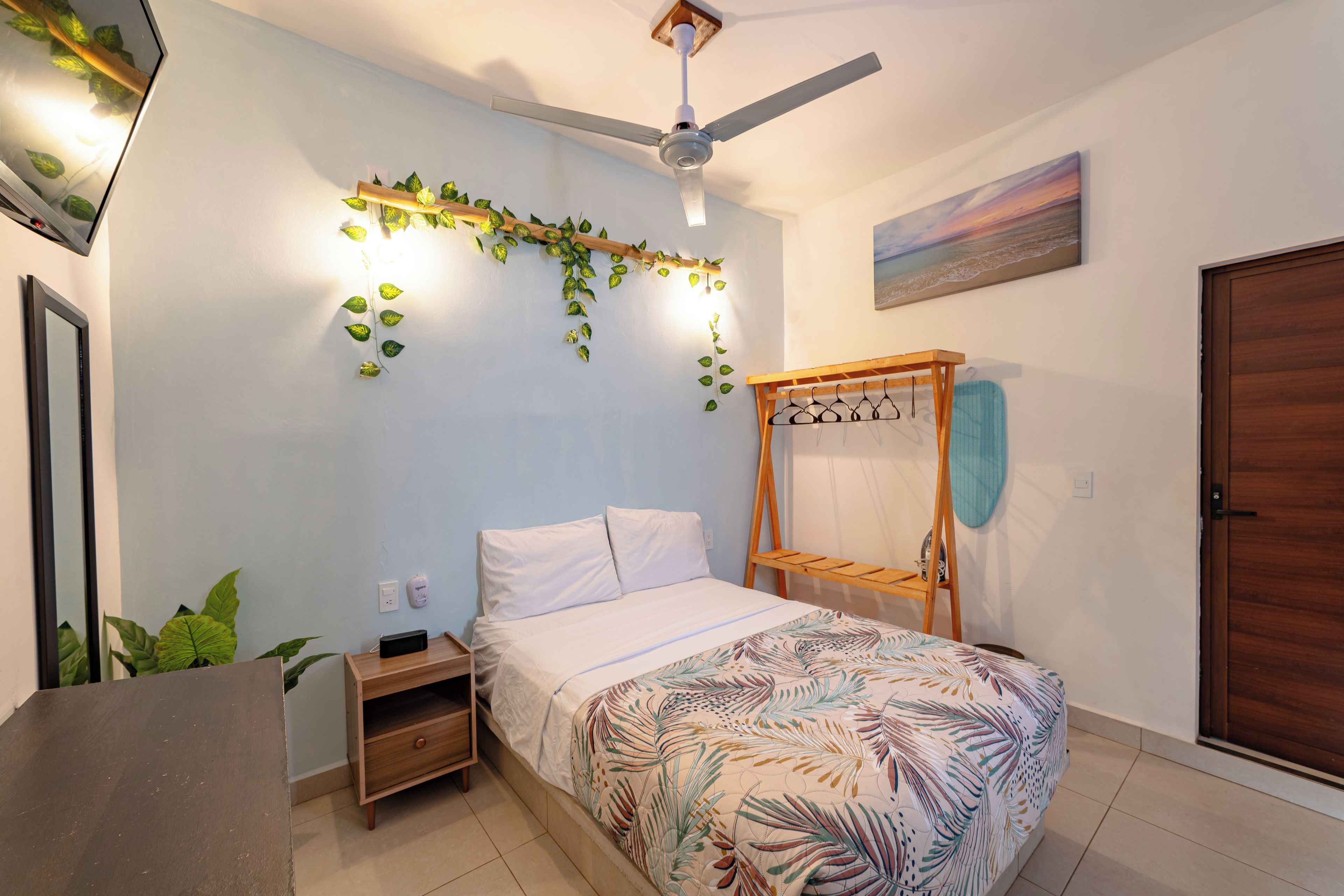 Une chambre à coucher avec un lit, un ventilateur de plafond, une étagère fixée au mur avec des plantes, un tableau et une porte en bois.