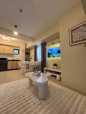 A modern living room with a flat-screen TV, a dining area with chairs, a kitchen with wooden cabinets, and a large window. - Cozy 1-bedroom condo in Serin East Tagaytay (Tagaytay City)