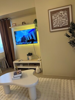 A living room with a flat-screen TV mounted on the wall, a white coffee table with books, a small white cabinet, and a framed abstract art piece. - Cozy 1-bedroom condo in Serin East Tagaytay (Tagaytay City)