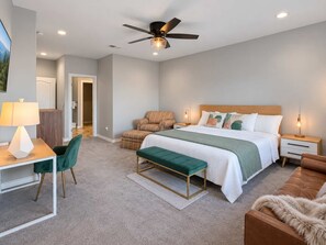 A modern bedroom with a bed, bedside tables, a desk, a chair, a bench, a ceiling fan, and a door leading to another room. - Spacious 4 Bedroom Mountain Home Near Lake and Golf (Ruidoso)