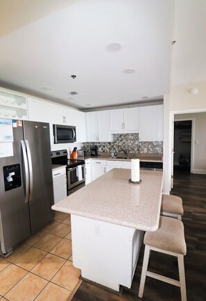A modern kitchen with white cabinetry, a stainless steel refrigerator, a microwave, and a large island with bar stools. - Emerald Escape - 3BR - Resort Pools & Bay Views (Panama City Beach)