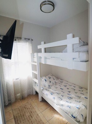 Habitación con literas, un televisor montado en la pared, una ventana con cortinas y piso de baldosas.