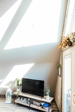 A modern living room with a flat-screen TV on a wooden stand, a 'hello' sign on the floor, and a large skylight. - Starlight - Pondside  Hot Tub - Game Room & Kayaks (Campton)