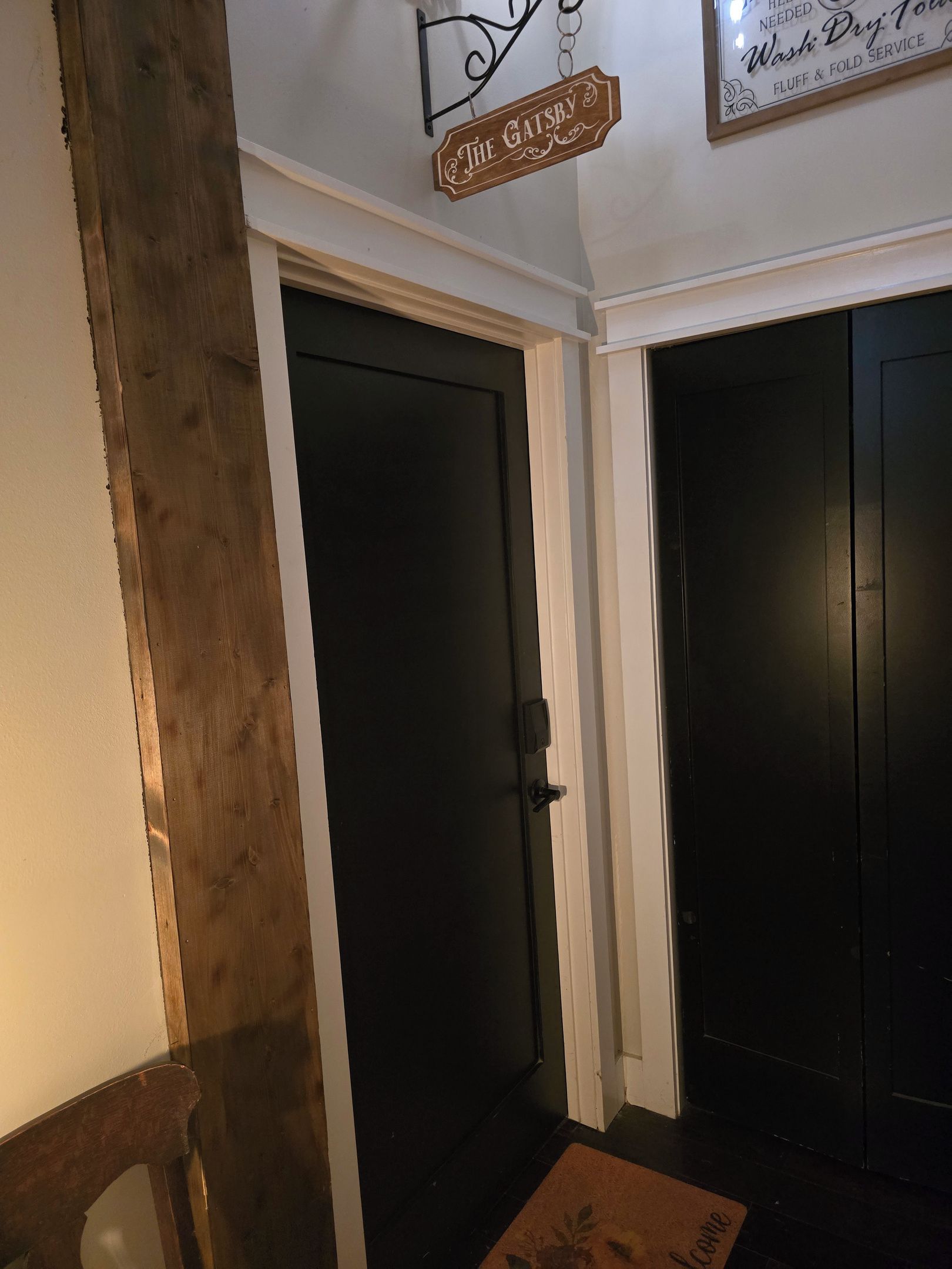 A hallway with two black doors, one with a wooden sign above it, and a welcome mat at the entrance.