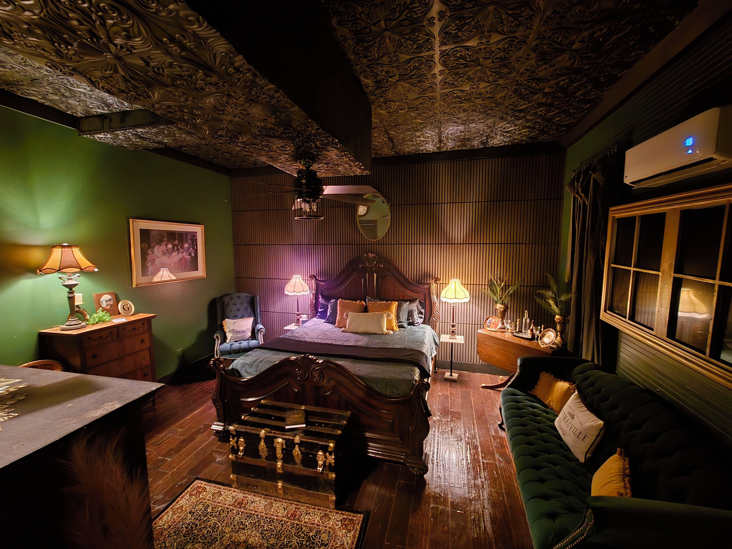 A room with a bed, a green sofa, a wooden dresser, a desk, and a ceiling with intricate designs.