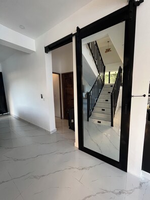 A spacious interior with a marble floor, a black-framed sliding door leading to a staircase, and a view of a second-floor balcony. - BellaHills by Ambar (San Juan)