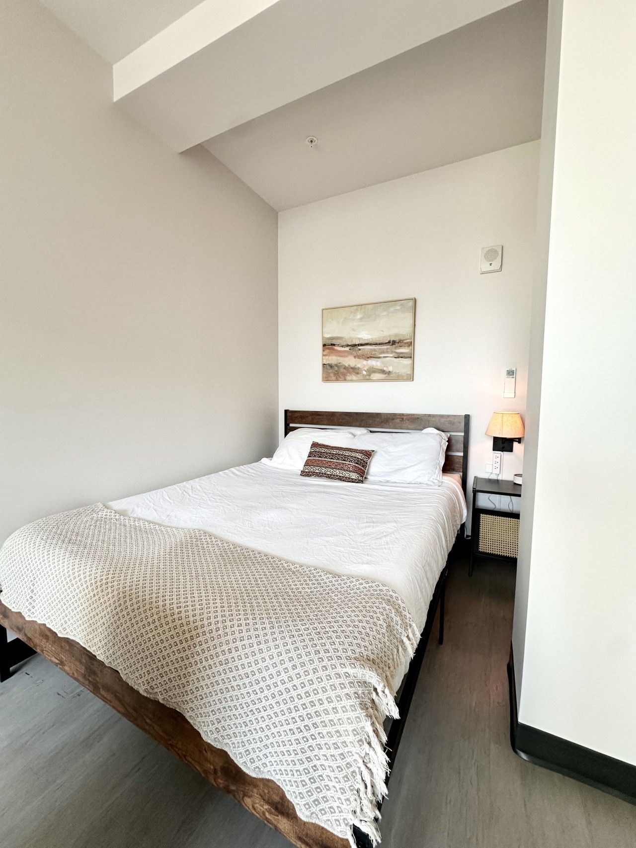A bedroom with a bed, a bedside table with a lamp, a wall-mounted light, and a painting.