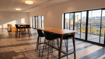A spacious room with a large table and chairs, large windows with a city view, and a high ceiling with recessed lighting.