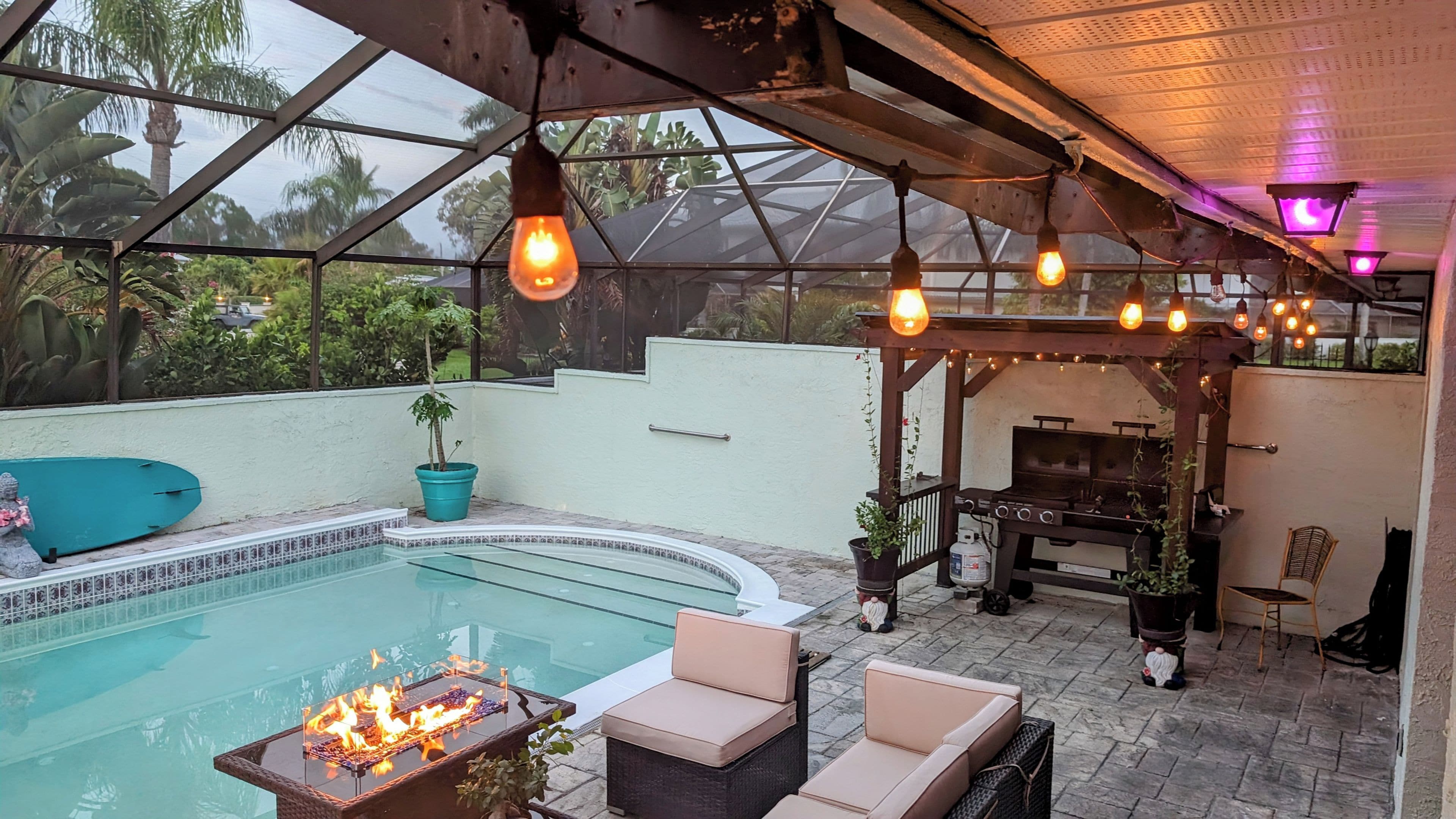 An outdoor pool area with a glass enclosure, lounge chairs, a barbecue grill, and hanging lights.
